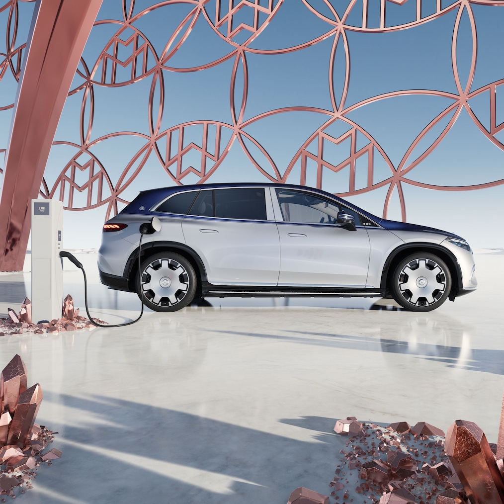 มุมมองด้านข้างของรถยนต์ SUV Mercedes-Maybach EQS ที่สถานีชาร์จไฟ พร้อมโลโก้แบรนด์ Maybach ที่พื้นหลัง