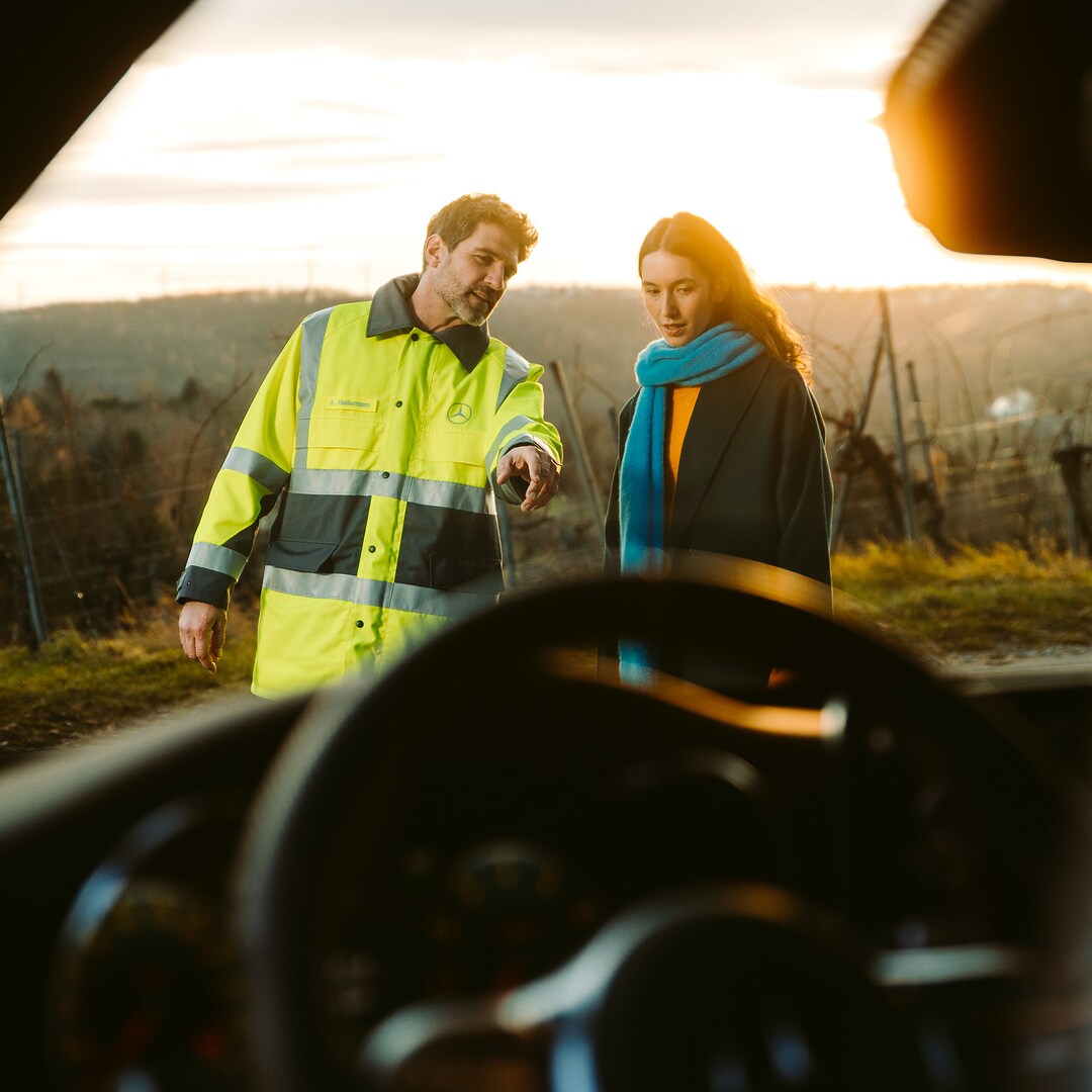 Through the windscreen of a Mercedes we see a service technician standing next to the driver in front of the vehicle and pointing at it to explain something.
