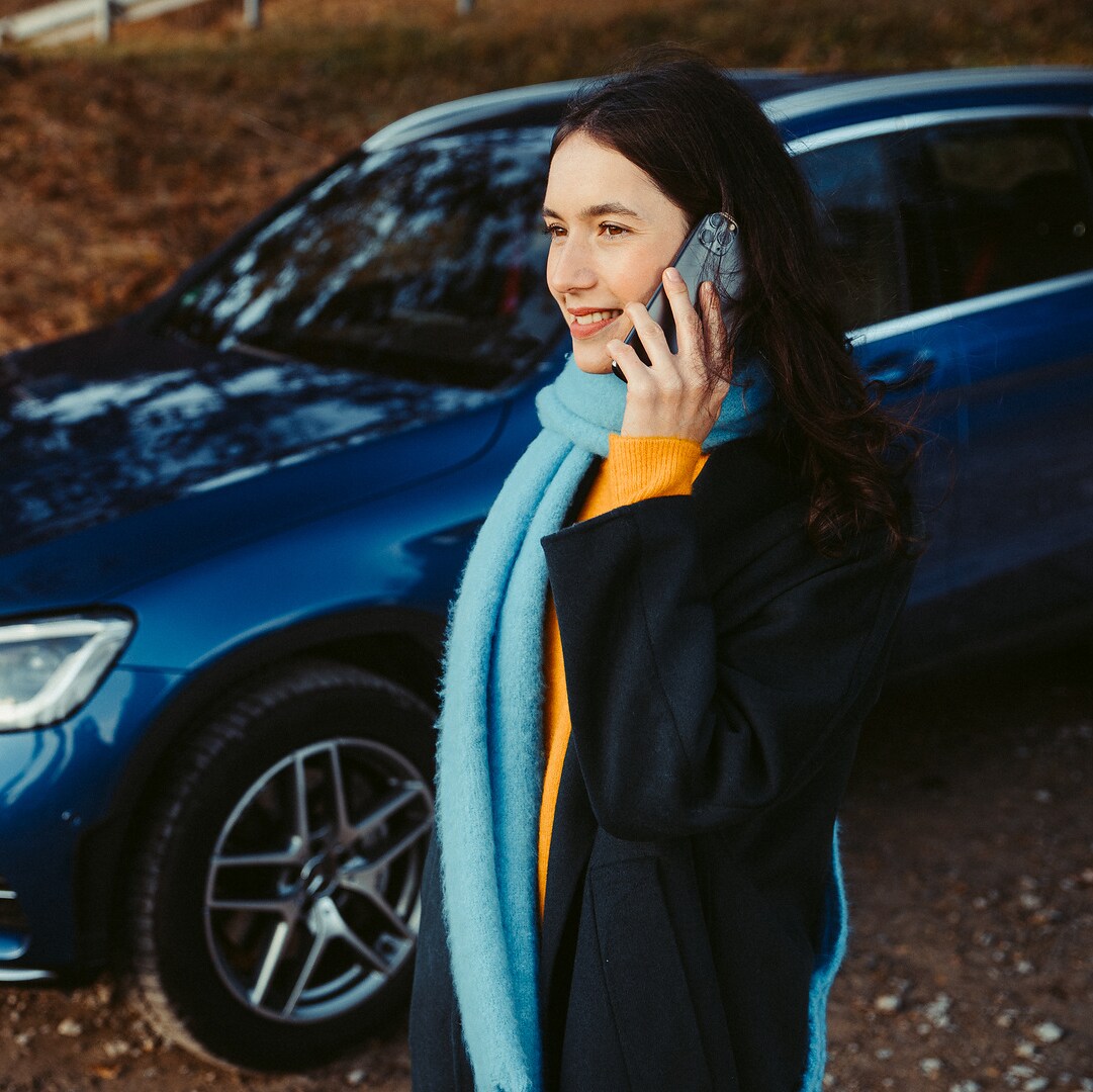 ผู้หญิงคนหนึ่งยืนพิงรถยนต์ Mercedes-Benz และจ้องสมาร์ทโฟนของเธอ