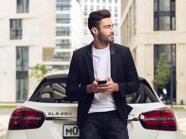 A man leans against his Mercedes-Benz GLA and uses the Mercedes me App on his smartphone and the Navigation Digital Extras to find out what the weather will be like at his destination.