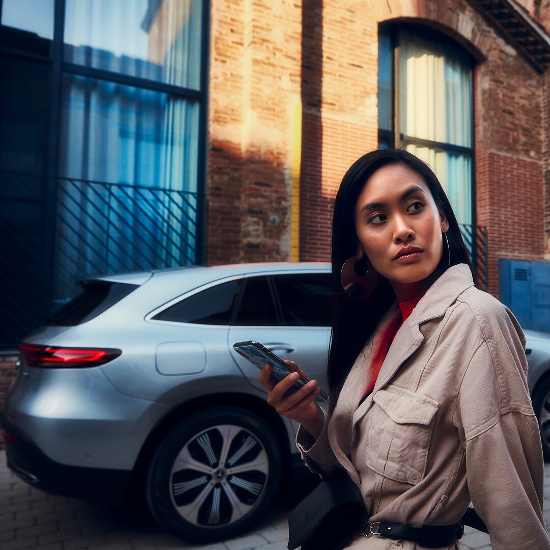 A lady standing in front of her Mercedes with a smartphone in her right hand.